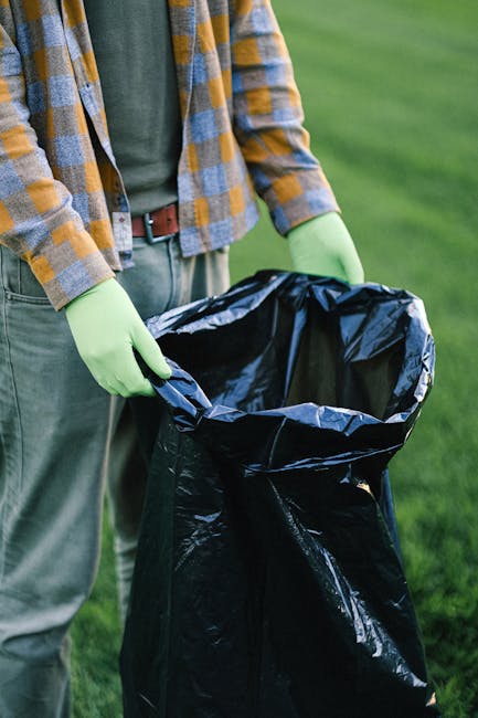 The image shows a person wearing a plaid shirt in shades of yellow, gray, and white, with a green long-sleeve garment underneath, standing outdoors on a grassy area during daylight. The individual is using bright green gloves to hold open a large black plastic rubbish bag, which appears partially filled and is positioned on the ground. The background features a well-maintained lawn with vibrant green grass, suggesting a garden or park setting. The scene emphasizes the act of waste collection or rubbish removal, aligning with services such as independent rubbish disposal or private waste handling provided by companies like House Clearance Ruislip. The lighting is natural, highlighting the textures of the fabrics, gloves, and plastic bag, conveying a professional and neutral tone suitable for informative content related to rubbish removal or clearance services in a residential or outdoor environment.