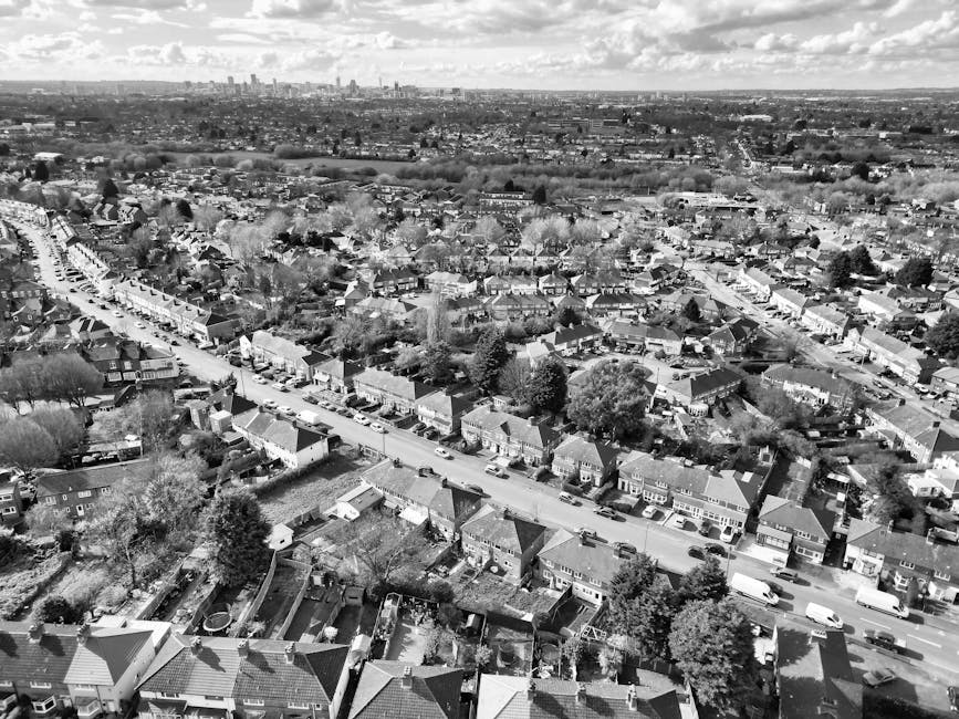 An aerial black-and-white photograph captures a densely populated residential neighbourhood with a mixture of detached, semi-detached, and terraced houses closely aligned along a curving main street. The houses display various roof styles, primarily pitched, with finishes including tiles and slate, and some rooftops featuring chimneys. The scene is filled with mature trees scattered throughout the area, creating a leafy environment, with many gardens visible behind fences or walls. In the foreground, residential backyards contain small sheds, patios, and garden furniture, with some yards showing accumulation of garden debris or clutter. The street is lined with parked cars on both sides, including sedans and small vans, and is illuminated by natural daylight under partly cloudy sky conditions. The distant horizon reveals a broader urban skyline with high-rise buildings, indicating a larger city nearby. This landscape, observed by House Clearance Ruislip, exemplifies typical suburban properties that may require private rubbish removal or on-site clearance for household waste disposal, separate from local authority collections.