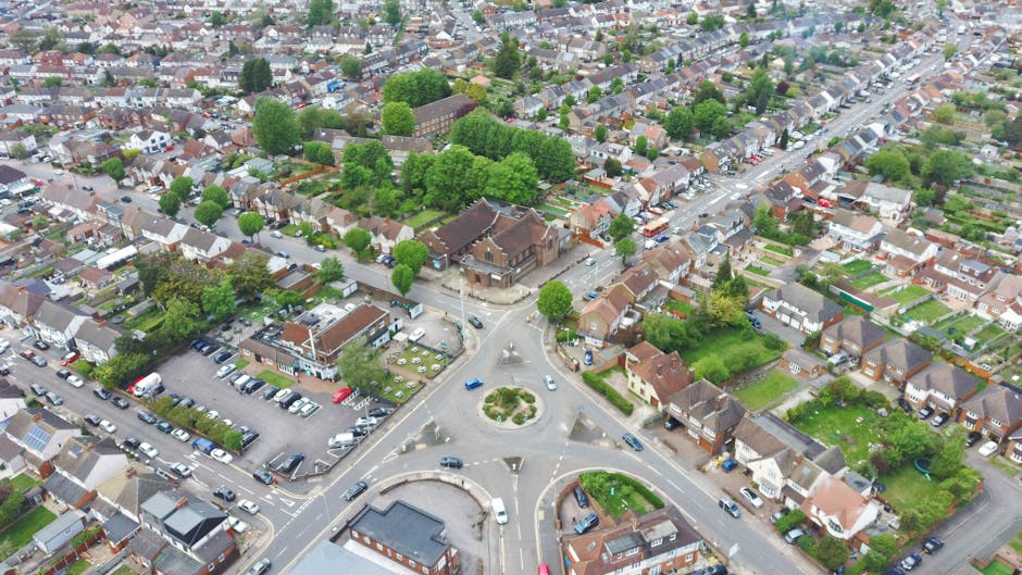 An aerial view of a residential neighbourhood featuring a roundabout with multiple roads extending from it. Surrounding the roundabout are small houses with pitched roofs, some with driveways and garages, and gardens with lawns and trees. The area is densely populated with closely spaced homes, many with green foliage and well-maintained yards. To the left of the roundabout, there is a parking lot filled with various parked cars, adjacent to a building with a flat roof and signage visible from above. The streets are lined with trees, and several vehicles are seen moving along the roads. In the background, more rows of houses and green spaces stretch into the distance, all under a slightly overcast sky. The scene provides a typical suburban setting that could be associated with private waste collection services, such as those offered by House Clearance Ruislip, and reflects an environment where onsite rubbish removal or alternative waste handling might be relevant.